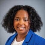 A smiling woman with curly black hair wears a blue blazer over a white shirt, posing in front of a gray background.