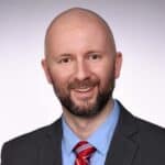 A bald man with a beard is smiling, dressed in a gray suit, blue shirt, and red striped tie, against a neutral background.