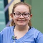 A female healthcare student wearing blue scrubs and glasses smiles for the camera.