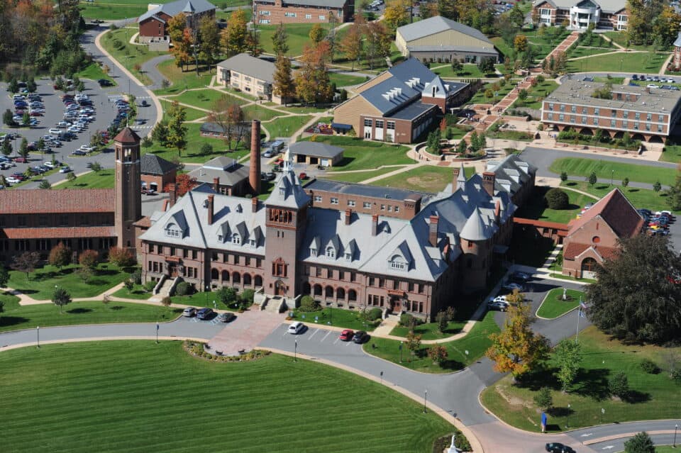 Aerial view of a large, historical-looking building with a clock tower, surrounded by smaller buildings and parking lots on a grassy campus. Paths connect the various structures.