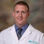 A person wearing a white lab coat, light blue shirt, and striped tie, posing for a photograph. The lab coat has a logo and the text "Conemaugh Health System.