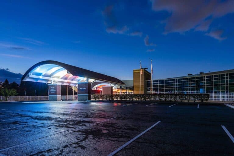 A modern building with a curved roof, illuminated at night with blue and white lights. The structure is adjacent to a large, empty parking lot under a dark, cloudy sky.